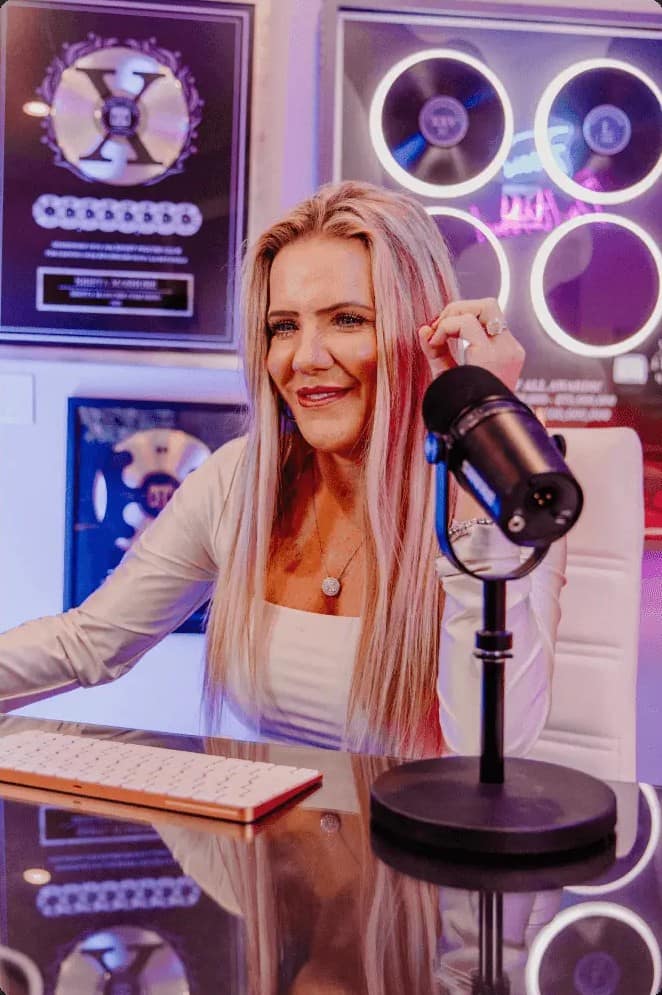 Krista Mashore at a microphone in her studio, with framed award plaques and record-style award discs mounted on the wall behind her.